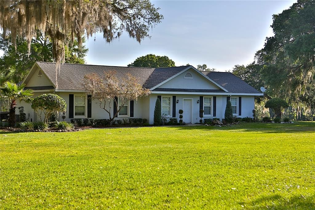 This is a front exterior view of a single-story home with a well-maintained lawn. The house features a gray roof, light-colored siding, and dark shutters. Landscaping includes trimmed bushes and trees, adding to the property's curb appeal.