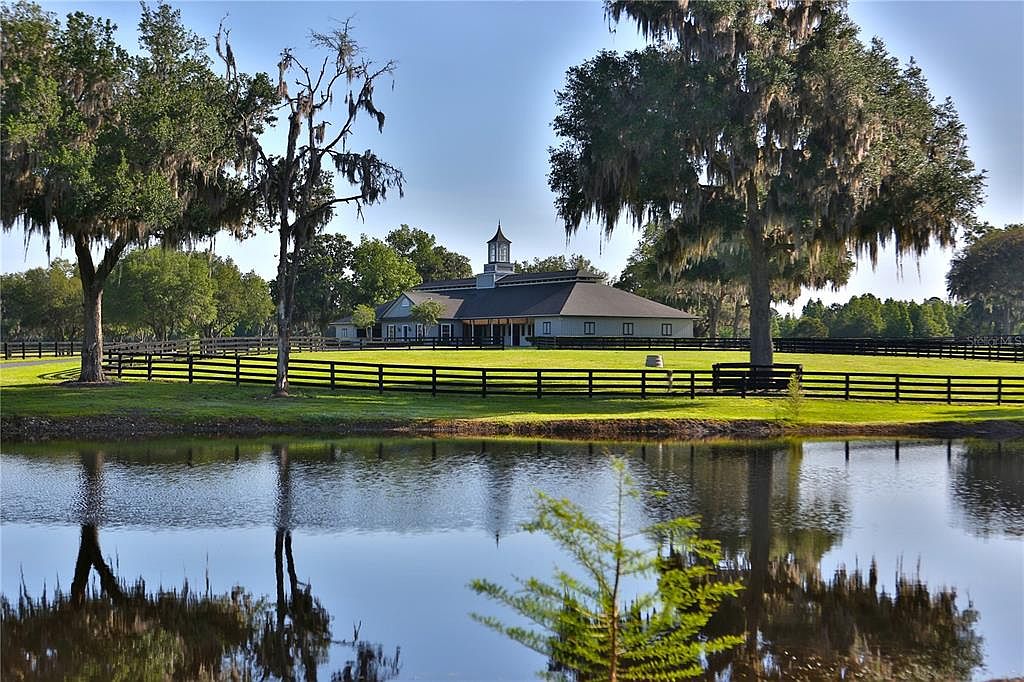 This exterior shot showcases a serene yard with a large pond reflecting the surrounding landscape. A black wooden fence encloses a lush green lawn, leading up to a building with a cupola. Mature trees with Spanish moss add to the property's charm, creating a picturesque and tranquil setting.