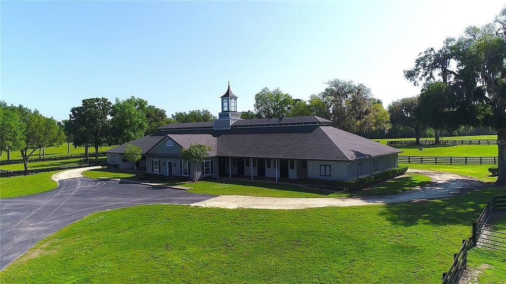 This aerial view showcases a large, well-maintained building with a dark roof and a central cupola. The surrounding landscape features lush green lawns, mature trees, and fenced pastures, creating a serene and spacious estate. A paved driveway leads to the building, enhancing its accessibility and curb appeal.