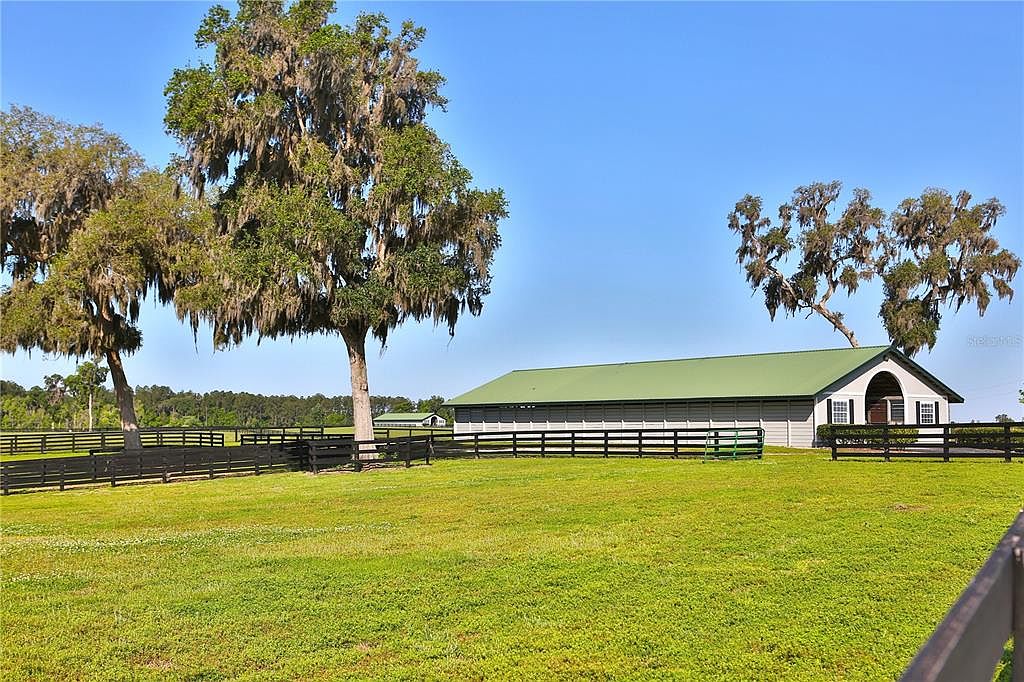 The image showcases a well-maintained yard with lush green grass and mature trees, some draped with Spanish moss. A long, light gray barn-like structure with a green roof is visible in the background, partially obscured by the trees. A black wooden fence runs along the perimeter of the property, adding a sense of enclosure and privacy.