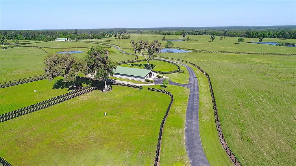 This aerial shot showcases a sprawling estate with lush green pastures, dark wooden fences, and a long driveway leading to a central building with a light green roof. Several ponds dot the landscape, adding to the property's serene and expansive feel. The image emphasizes the property's size, privacy, and well-maintained grounds, making it ideal for equestrian or agricultural pursuits.