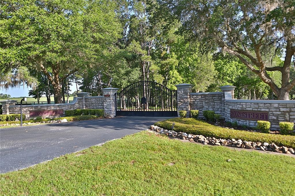 The image showcases an impressive gated entryway to a property, featuring stone pillars and walls with the name "STONESTREET" displayed. A black wrought-iron gate provides security and elegance, while lush greenery and mature trees create a serene and private atmosphere. The paved driveway leads into the estate, suggesting a well-maintained and prestigious residence.