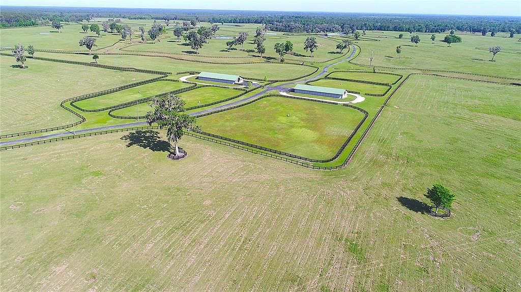 This aerial shot showcases a sprawling estate with lush green pastures, fenced paddocks, and well-maintained landscaping. Two large, modern barns or stables are centrally located, connected by a paved driveway. The property exudes a sense of luxury and spaciousness, ideal for equestrian pursuits or a private retreat.