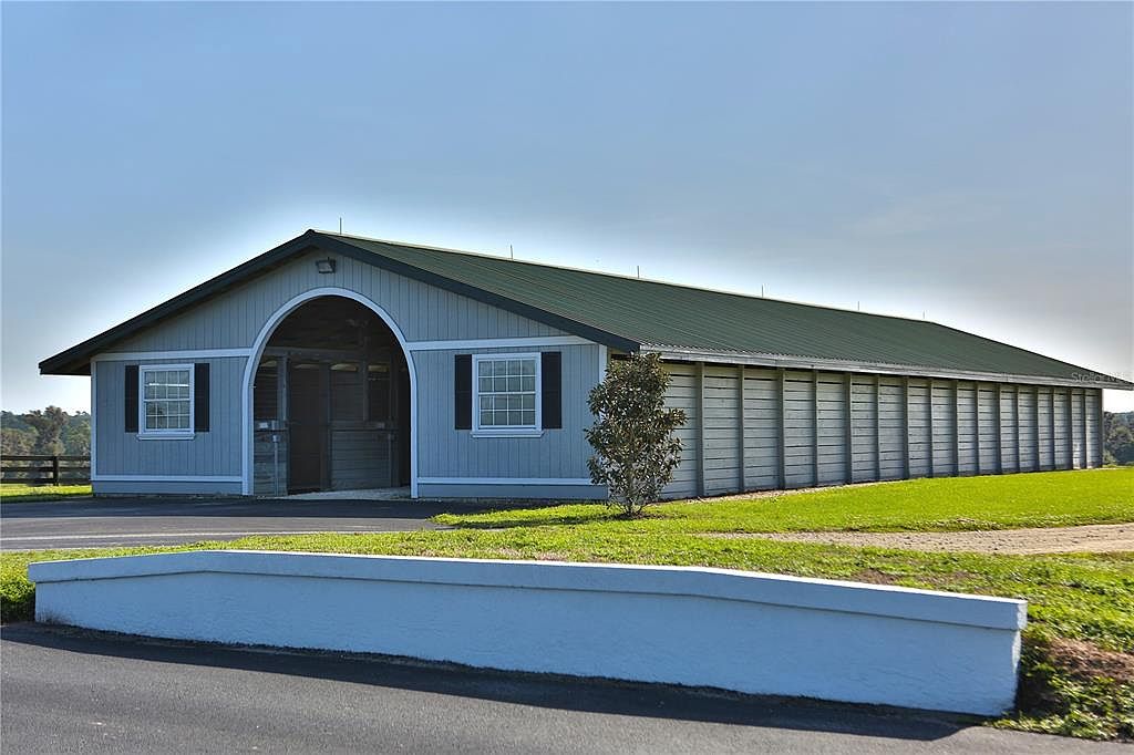 The image showcases the front exterior of a long, single-story barn-like structure with a gray facade and a dark green roof. The building features an arched entryway and symmetrically placed windows with dark shutters. A well-maintained lawn surrounds the building, and a low white wall runs along the front, adding to the property's curb appeal.