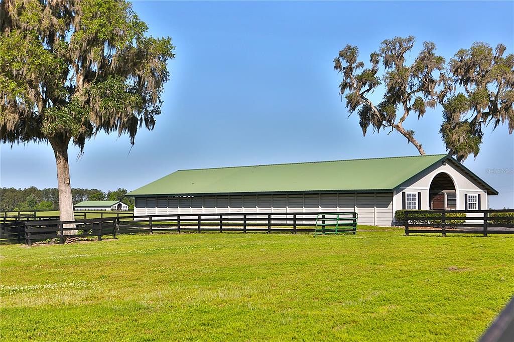The image showcases a well-maintained yard with a large green lawn and a long, light gray barn-like structure with a green roof. A black wooden fence runs along the perimeter, adding a touch of rustic charm. Mature trees with Spanish moss frame the scene, creating a serene and picturesque setting.