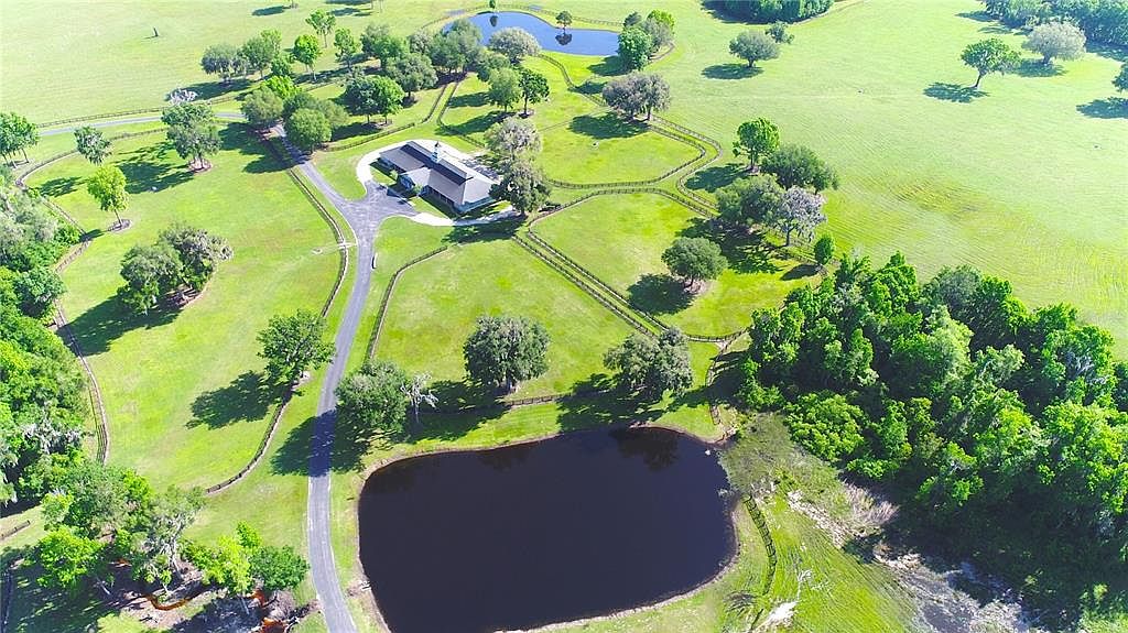 This aerial shot showcases a sprawling estate with a modern home, multiple ponds, and fenced pastures. The property is surrounded by lush green lawns and mature trees, creating a serene and private setting. A long driveway leads to the house, emphasizing the property's grandeur and seclusion.