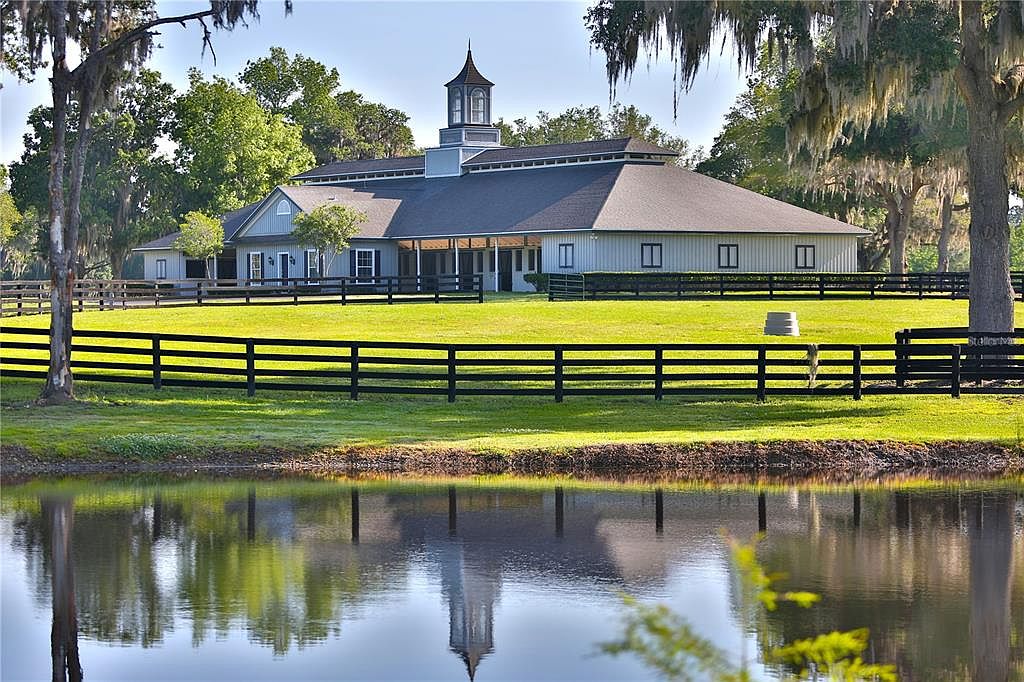 This image showcases the front exterior of a large, well-maintained building, possibly a barn or estate house, with a distinctive cupola. The property features a lush green lawn enclosed by a black wooden fence, and a serene pond reflects the building's facade, creating a picturesque scene. Mature trees with Spanish moss add to the property's charm and established feel.
