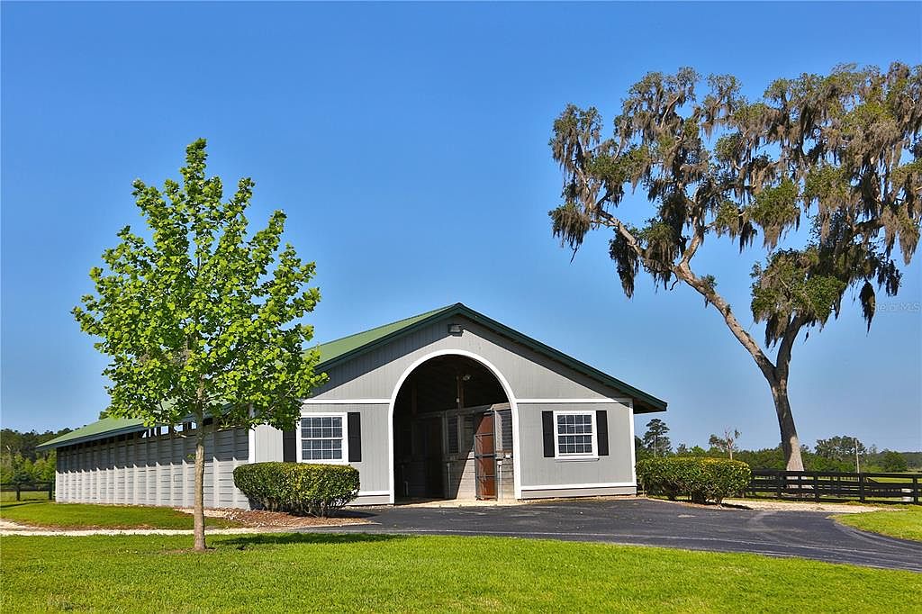 The image showcases the front view of a well-maintained stable or barn. The building features a gray exterior with a green roof, complemented by black window shutters. A neatly manicured lawn and mature trees frame the structure, creating a serene and inviting atmosphere, suggesting a property focused on equestrian activities or rural living.