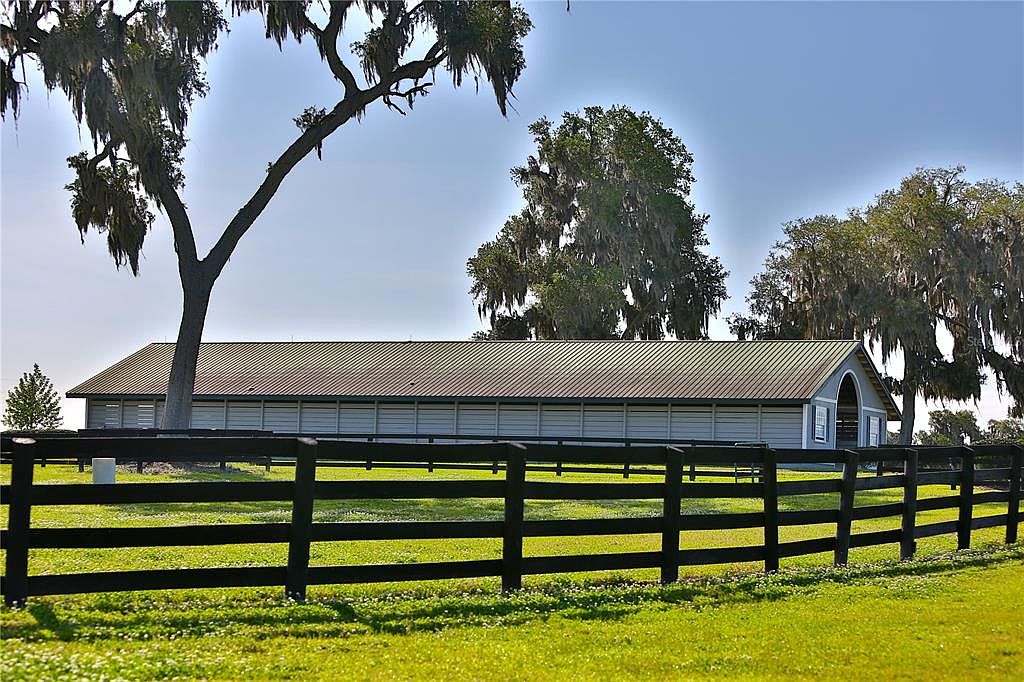 The image showcases a well-maintained yard with a long, low building in the background, possibly a barn or stable. A black wooden fence runs across the foreground, defining the property line. The lush green grass and mature trees with Spanish moss create a serene, pastoral setting, suggesting a spacious and private estate.
