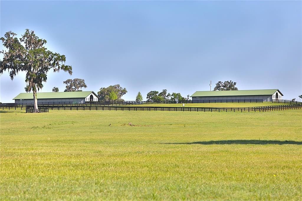 This image showcases a sprawling green field with two large, gray barns in the background, each featuring a green roof and a simple, functional design. A black wooden fence runs along the perimeter of the field, adding a touch of rustic charm and defining the property line. The clear blue sky above enhances the sense of openness and tranquility, making it an ideal setting for a rural estate.