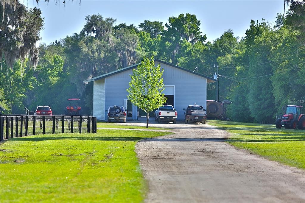 The image showcases a well-maintained yard with a large, light-blue barn-like structure in the background. A gravel driveway leads up to the building, flanked by green lawns and a small tree in the center. Several vehicles and farm equipment are parked near the building, suggesting a rural or agricultural property.
