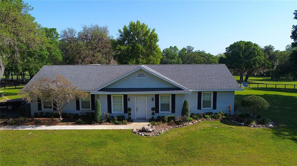 This is a front exterior view of a single-story house with a gray roof and light blue siding. The house features a symmetrical design with a central front door, flanked by windows with black shutters. A well-manicured lawn and landscaping add to the property's curb appeal, creating a welcoming and tidy impression.