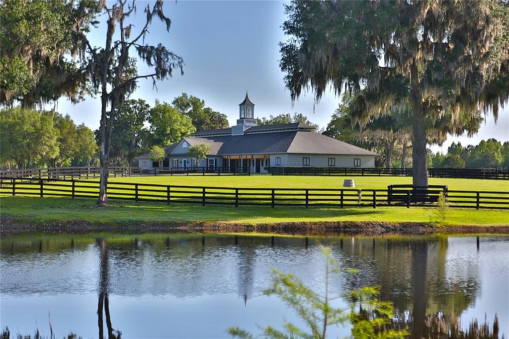 This image showcases the front exterior of a grand estate, featuring a large building with a distinctive cupola and a symmetrical design. A well-maintained lawn stretches towards a black wooden fence, beyond which lies a serene pond reflecting the building and sky. Mature trees frame the scene, adding to the property's established and picturesque appeal.
