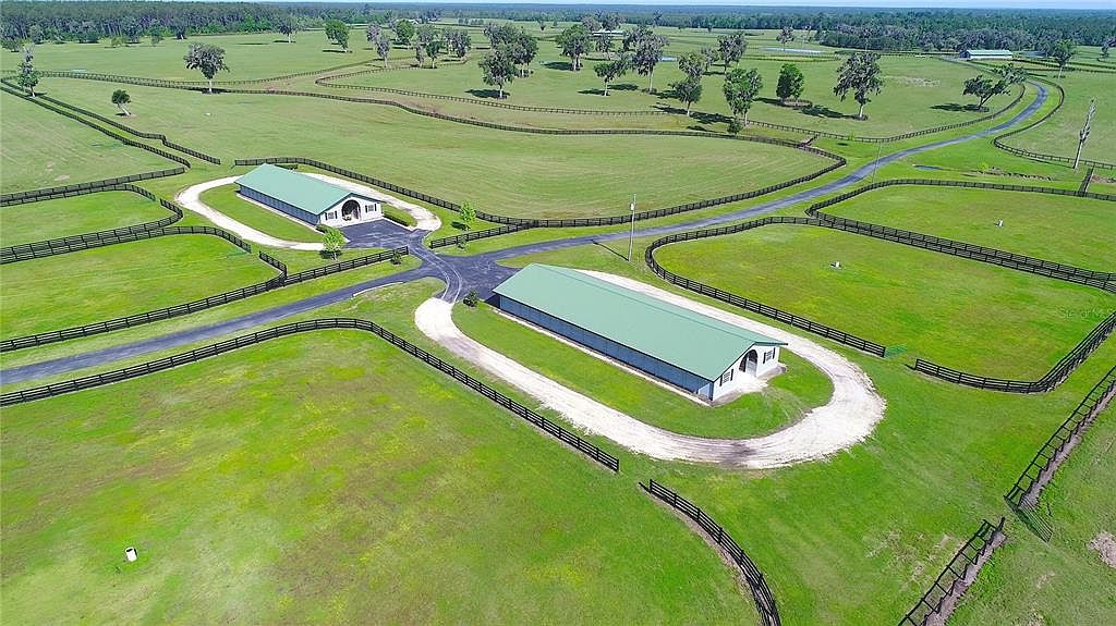 This aerial shot showcases a sprawling estate with lush green pastures, dark wooden fences, and two large buildings with green roofs. A network of paved and gravel driveways connects the buildings and meanders through the property, highlighting the expansive and well-maintained grounds. The perspective gives a sense of the property's scale and privacy.