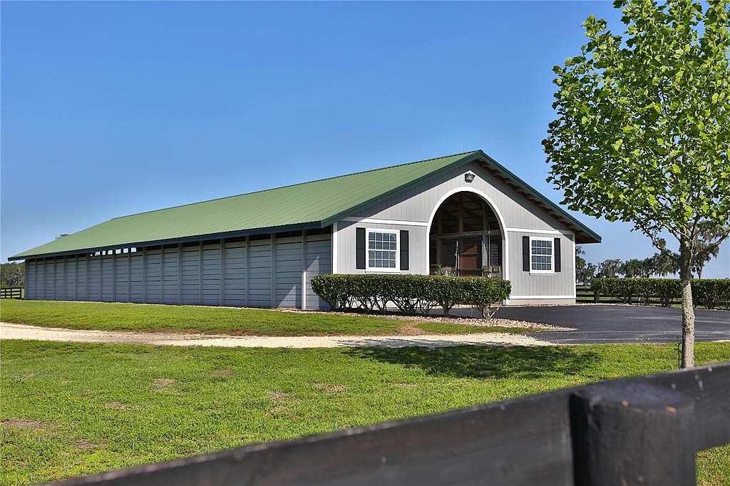 The image showcases the front view of a large, well-maintained stable or barn. It features a light gray exterior with a green metal roof, complemented by neatly trimmed hedges and a paved driveway. The structure includes multiple stalls or sections, suggesting ample space for livestock or storage, and the overall impression is one of functionality and quality.