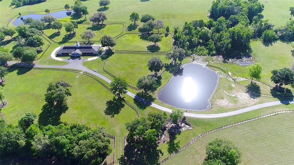 This aerial view showcases a sprawling estate with lush green lawns, mature trees, and a central building with a dark roof and light-colored facade. A network of fences and roads defines the property's boundaries, while ponds add to the scenic landscape. The overall impression is one of tranquility and spaciousness.