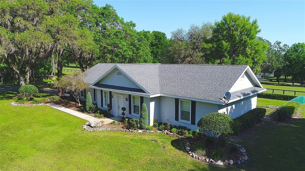 This is an aerial view of a single-story home with a gray roof, white exterior, and dark shutters. The house is surrounded by a well-manicured lawn and mature trees, creating a serene and private setting. A paved walkway leads to the front entrance, and a fence encloses part of the property.