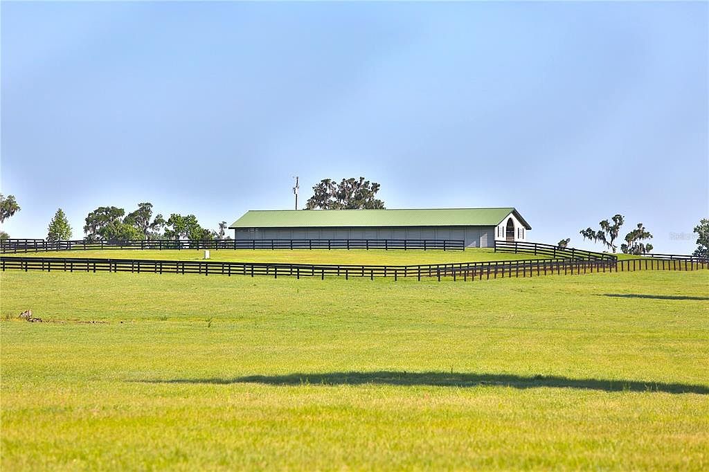 The image showcases a vast, well-maintained green field leading up to a large barn-like structure with a green roof. A dark wooden fence runs along the perimeter of the property, adding a touch of rustic charm. The clear blue sky provides a serene backdrop, emphasizing the spaciousness and tranquility of the estate.