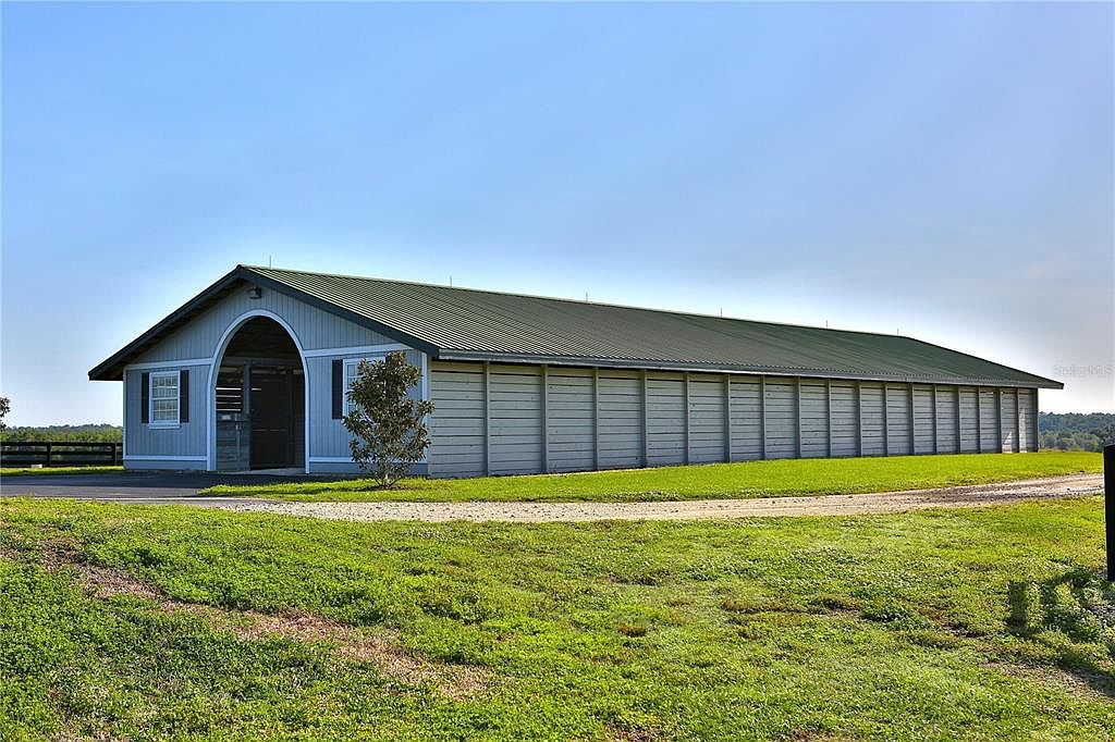 The image showcases the side view of a long, single-story barn or stable building with a green metal roof and light gray siding. The structure features a prominent arched entryway on one end and a series of sliding doors along its length. The surrounding landscape includes a well-maintained lawn and a gravel driveway, suggesting a rural or agricultural setting.