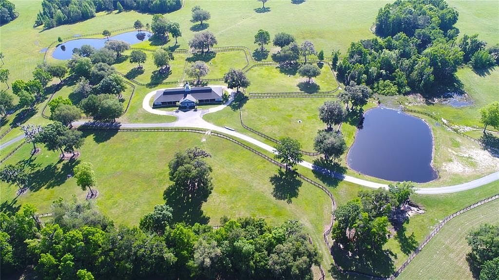 This aerial shot showcases a sprawling estate with lush green pastures, mature trees, and multiple ponds. A long driveway leads to a central building with a dark roof, surrounded by fenced enclosures. The property exudes a sense of privacy and tranquility, ideal for equestrian or agricultural pursuits.
