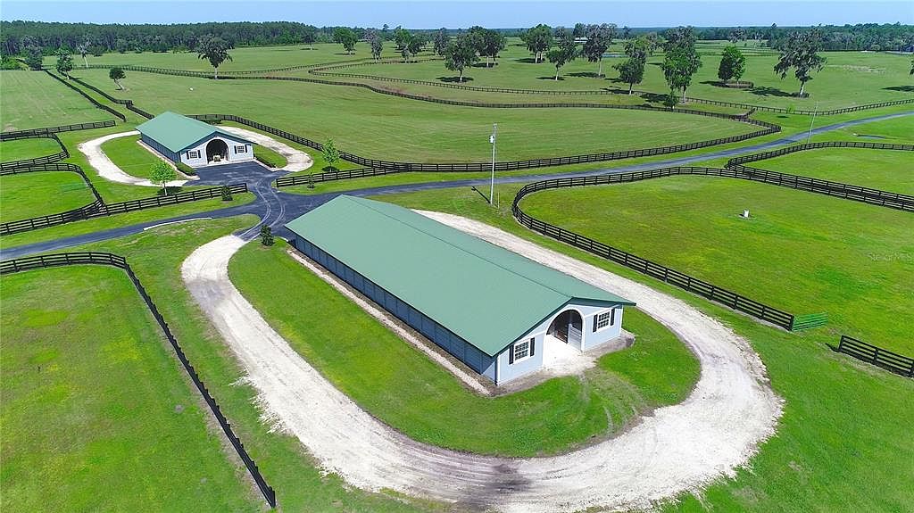 This aerial shot showcases a sprawling estate with lush green pastures and dark wooden fencing. Two large, light blue buildings with green roofs are prominently featured, each surrounded by gravel driveways. The property exudes a sense of luxury and spaciousness, ideal for equestrian or agricultural pursuits.