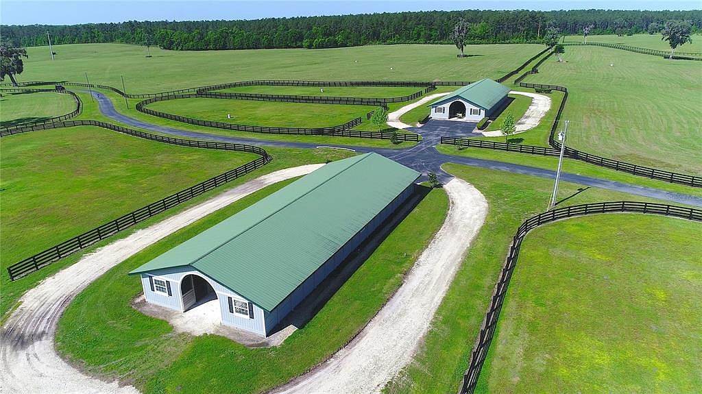 This aerial shot showcases a sprawling estate with well-maintained green pastures, dark wooden fences, and two large barns with light green roofs. A network of gravel driveways connects the buildings, creating a sense of organized luxury. The property is surrounded by lush trees, offering privacy and a serene atmosphere.