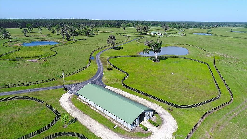 This aerial shot showcases a sprawling estate with lush green pastures, a large barn-like structure with a green roof, and several ponds. The property is enclosed by dark wooden fences, creating a picturesque and private setting. A long driveway leads to the building, emphasizing the scale and grandeur of the estate.