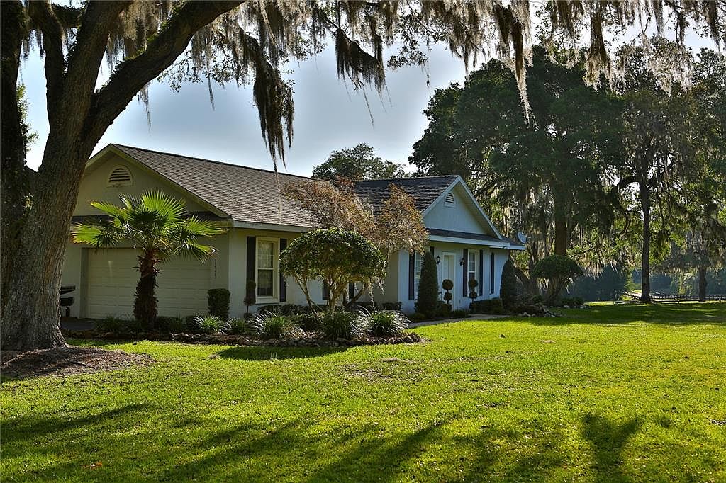 This is a front view of a single-story house with a well-maintained lawn. The house features a light-colored exterior, a dark roof, and dark shutters. Mature trees, some with Spanish moss, frame the house, adding to the property's charm and curb appeal. The perspective is from the front yard, showcasing the house and its landscaping.