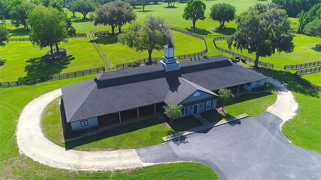 This aerial view showcases a sprawling estate with a large, well-maintained building featuring a dark roof and light blue accents. The property includes extensive green pastures enclosed by dark wooden fences, mature trees dotting the landscape, and winding gravel and paved driveways leading to the building. The overall impression is one of a luxurious and private rural retreat.