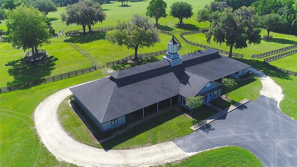 This aerial view showcases a sprawling estate with a large, well-maintained building featuring a dark roof and a central cupola. The property includes extensive green lawns, mature trees, and fenced pastures, creating a picturesque and serene setting. A circular driveway leads to the building, and a paved area provides additional parking.