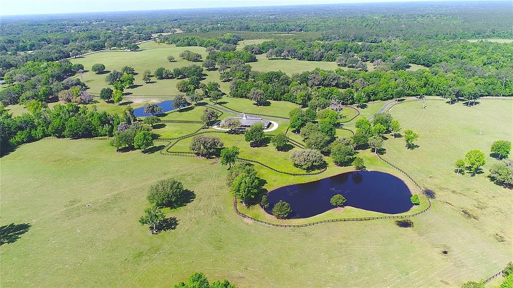 This aerial view showcases a sprawling estate with lush green pastures, mature trees, and two serene ponds. A well-maintained home sits nestled among the landscape, accessed by a winding driveway. The property is enclosed by fencing, adding to its privacy and charm.