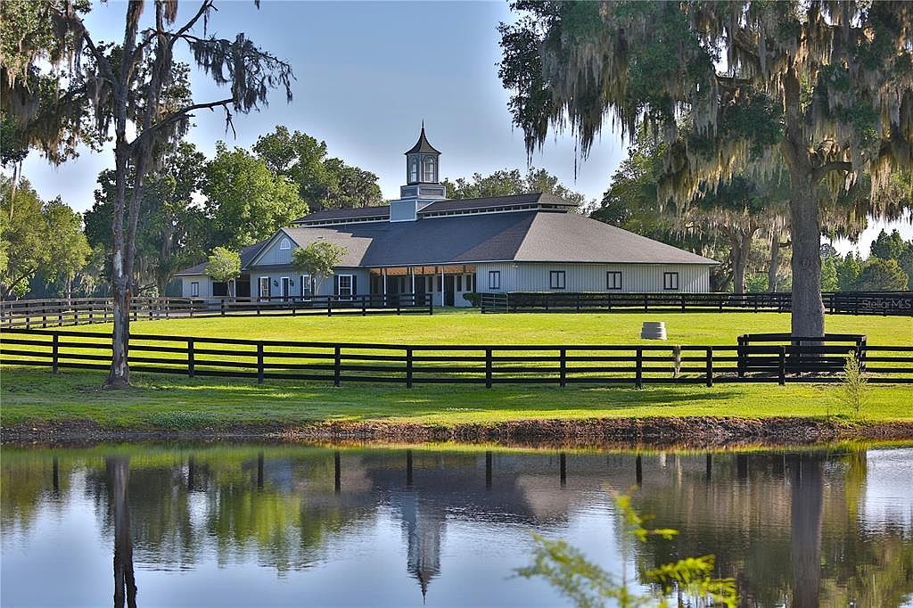This image showcases a large, well-maintained estate with a sprawling lawn and a sizable building, possibly a barn or stable, featuring a distinctive cupola. A black wooden fence surrounds the property, and a tranquil pond in the foreground reflects the building and sky, adding to the serene and picturesque setting. The presence of mature trees draped with Spanish moss enhances the property's charm and establishes a sense of timeless elegance.