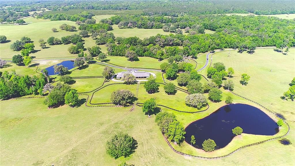 This aerial view showcases a sprawling estate with lush green pastures, mature trees, and two serene ponds. A well-maintained home sits at the center, accessed by a winding driveway, and the property is enclosed by elegant fencing. The overall impression is one of tranquility and luxury.