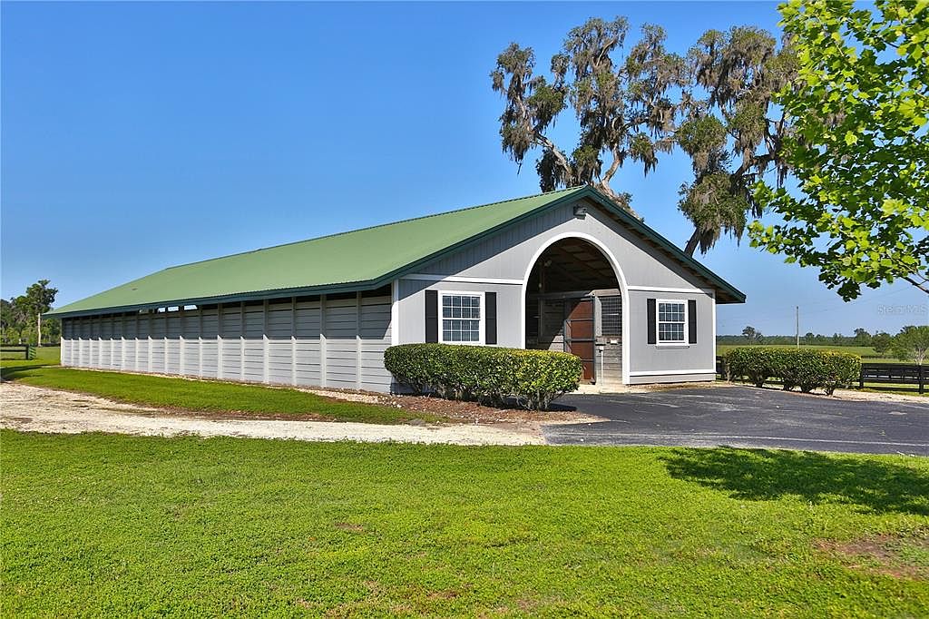 The image showcases the side view of a well-maintained stable or barn with a green roof and light gray siding. The structure features an arched entryway and several windows with dark shutters. Lush green grass and manicured bushes surround the building, creating a serene and picturesque setting.
