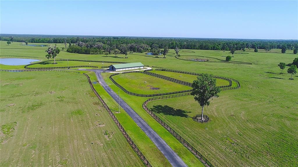 This aerial shot showcases a sprawling estate with lush green pastures, a long driveway leading to a central building with a light green roof, and fenced enclosures. Several ponds are visible in the distance, and the property is bordered by a dense treeline, creating a sense of privacy and seclusion. The view emphasizes the vastness and tranquility of the land.