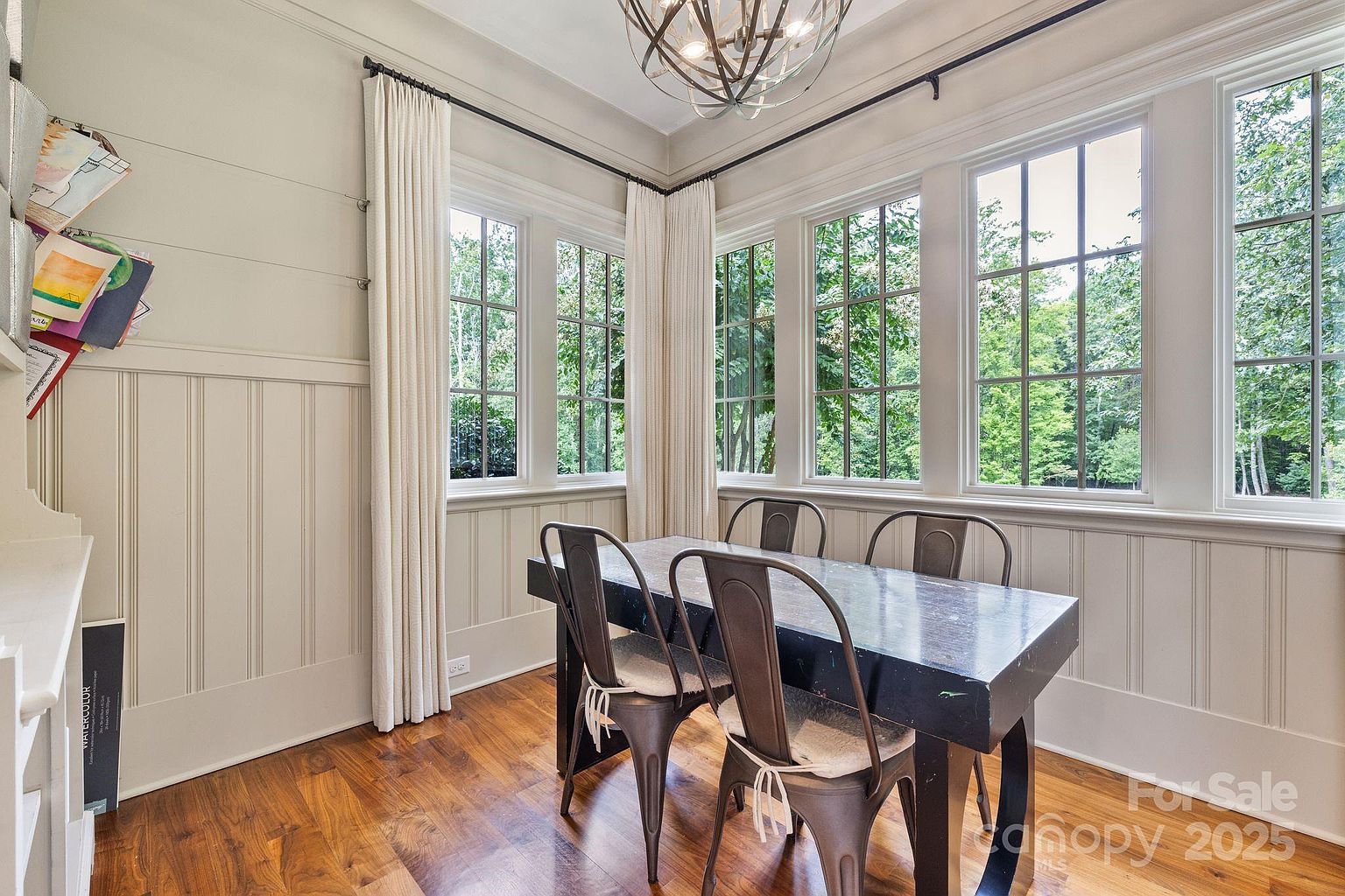This bright dining area features a dark rectangular table surrounded by four industrial-style metal chairs. The room is framed by large windows that offer a view of the surrounding trees, complemented by floor-length curtains and wainscoting on the lower walls. A decorative chandelier hangs above the table, creating a warm and inviting atmosphere for meals.