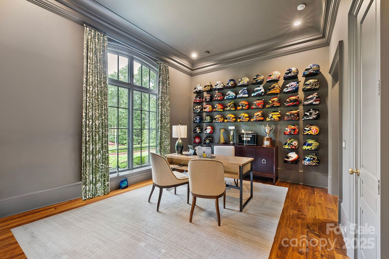 This sophisticated home office features a striking display wall showcasing a large collection of racing helmets arranged in a grid. The room is anchored by a wooden desk with two upholstered chairs, set against a backdrop of dark gray walls and tall, arched windows dressed in patterned green curtains. The space is finished with warm wood flooring and a neutral area rug, creating a professional yet personalized atmosphere.