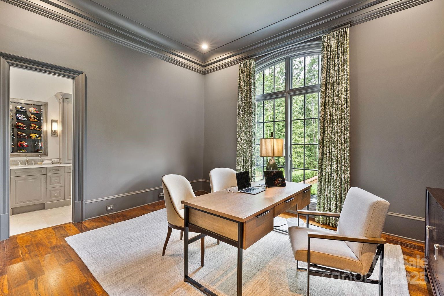This sophisticated home office features dark gray walls, crown molding, and rich wood flooring topped with a large area rug. A central wooden desk is positioned in front of a large, arched window that provides ample natural light and a view of the surrounding greenery. The room is furnished with three chairs and includes a doorway leading to an adjacent space displaying a collection of racing helmets.