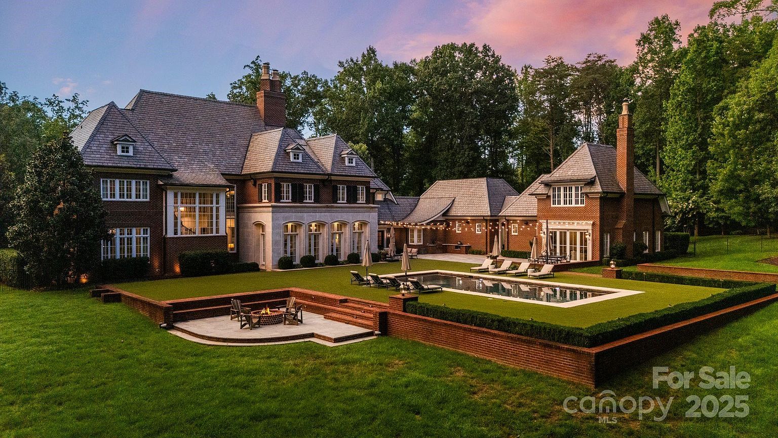 This expansive rear view of a grand brick estate showcases a sophisticated architectural design with multiple gables and a slate roof. The property features a meticulously landscaped backyard, a rectangular swimming pool surrounded by lounge chairs, and a sunken stone fire pit area. String lights draped across the patio area create an inviting atmosphere, while the surrounding mature trees provide a sense of privacy and seclusion.