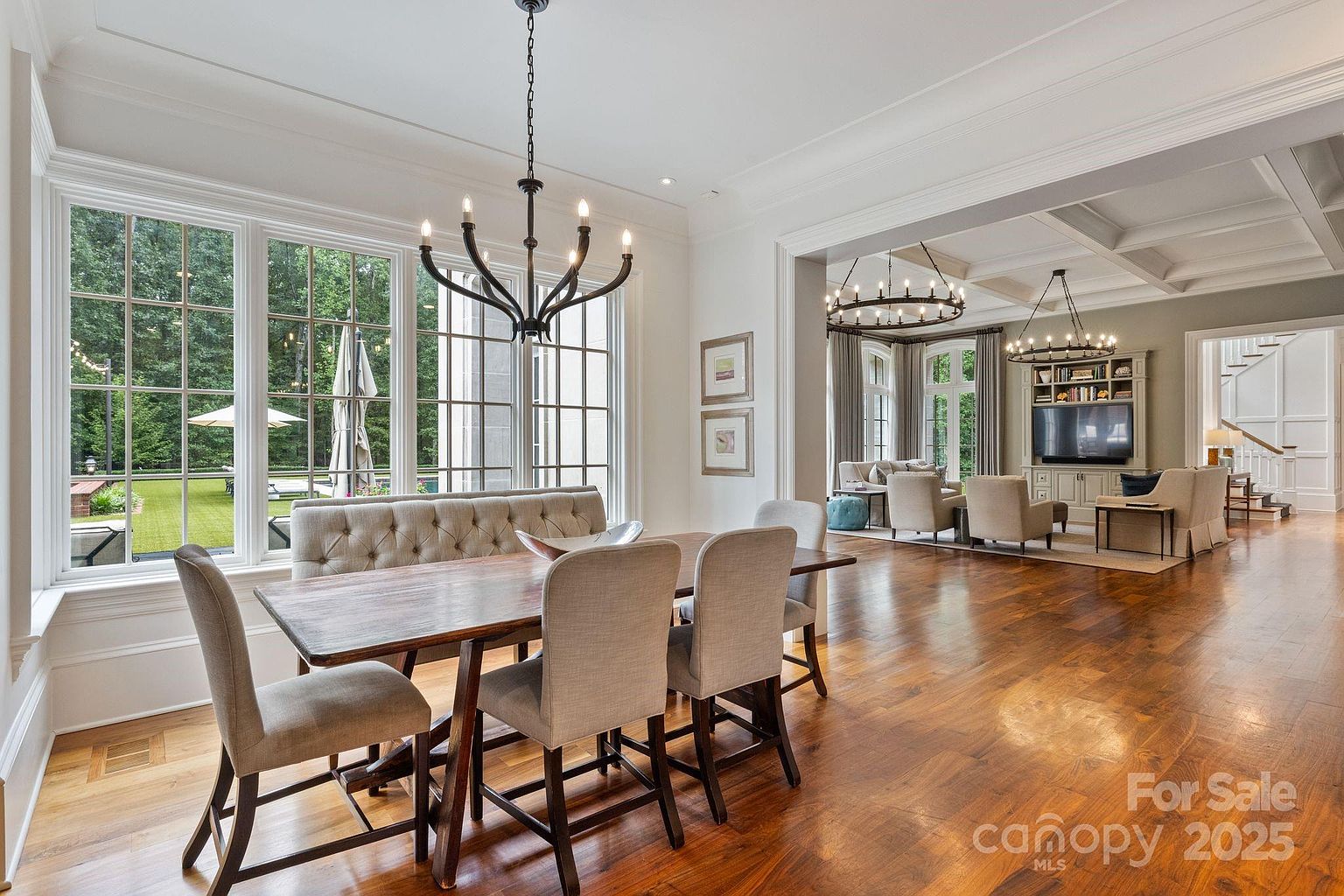 This elegant dining area features a wooden table paired with a tufted upholstered bench and four matching chairs, all illuminated by a dark metal chandelier. Large windows provide ample natural light and a view of the lush backyard, while the open floor plan flows seamlessly into a spacious living room with a built-in media center. The room is finished with warm-toned hardwood flooring and neutral wall colors, creating a sophisticated and inviting atmosphere.