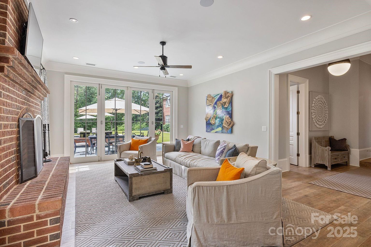 This bright and inviting living room features a neutral color palette with comfortable seating arranged around a central coffee table. A tall brick chimney breast anchors the space, while large glass doors provide a seamless transition to the outdoor patio area. The room is accented with colorful throw pillows and abstract wall art, creating a warm and sophisticated atmosphere.