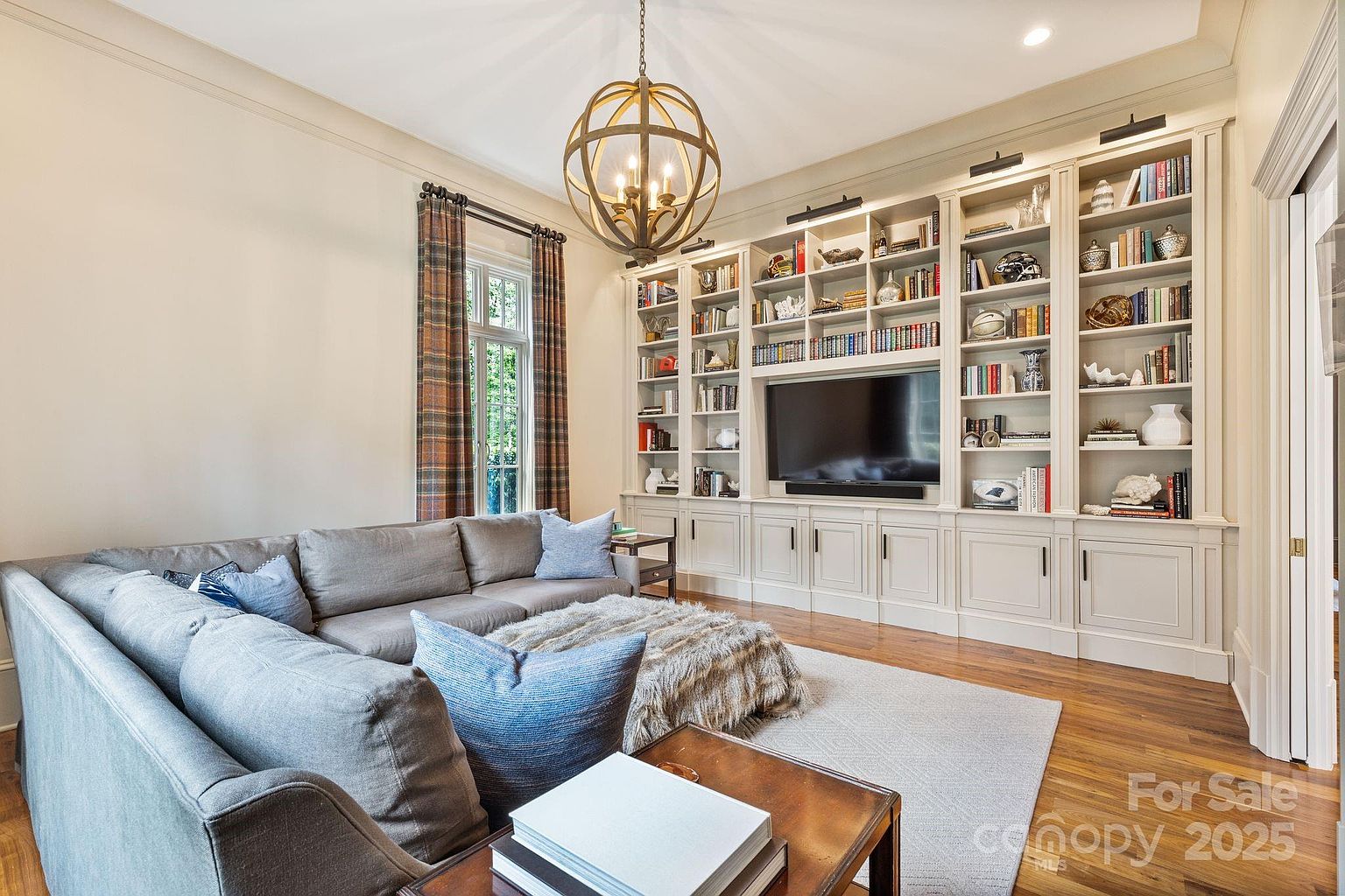 This inviting living room features a large, plush gray sectional sofa arranged around a textured ottoman, creating a cozy seating area. A prominent built-in wall unit with extensive shelving and cabinetry surrounds a central television, providing ample storage and display space. The room is illuminated by a striking spherical chandelier and natural light streaming through tall windows dressed in plaid curtains, all set against warm wood flooring.