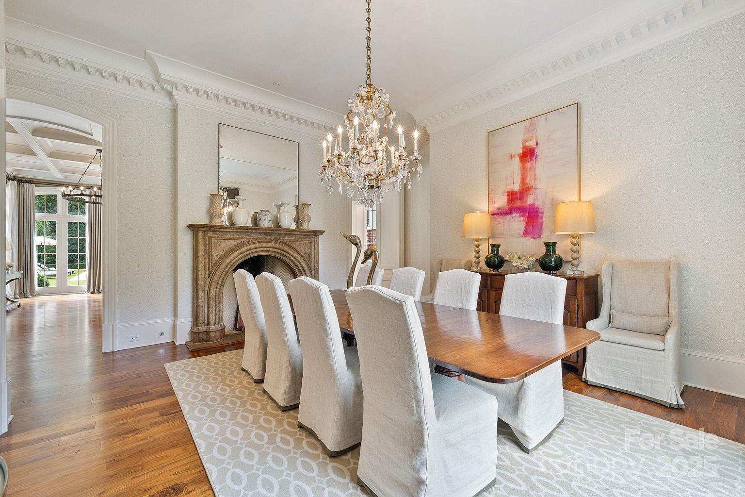This elegant dining room features a long wooden table surrounded by eight slipcovered chairs, set atop a patterned area rug. A prominent stone fireplace with a mirror above serves as the focal point, complemented by a crystal chandelier and a large abstract painting on the adjacent wall. The room is framed by intricate crown molding and opens into a bright hallway, creating a sophisticated and inviting atmosphere.