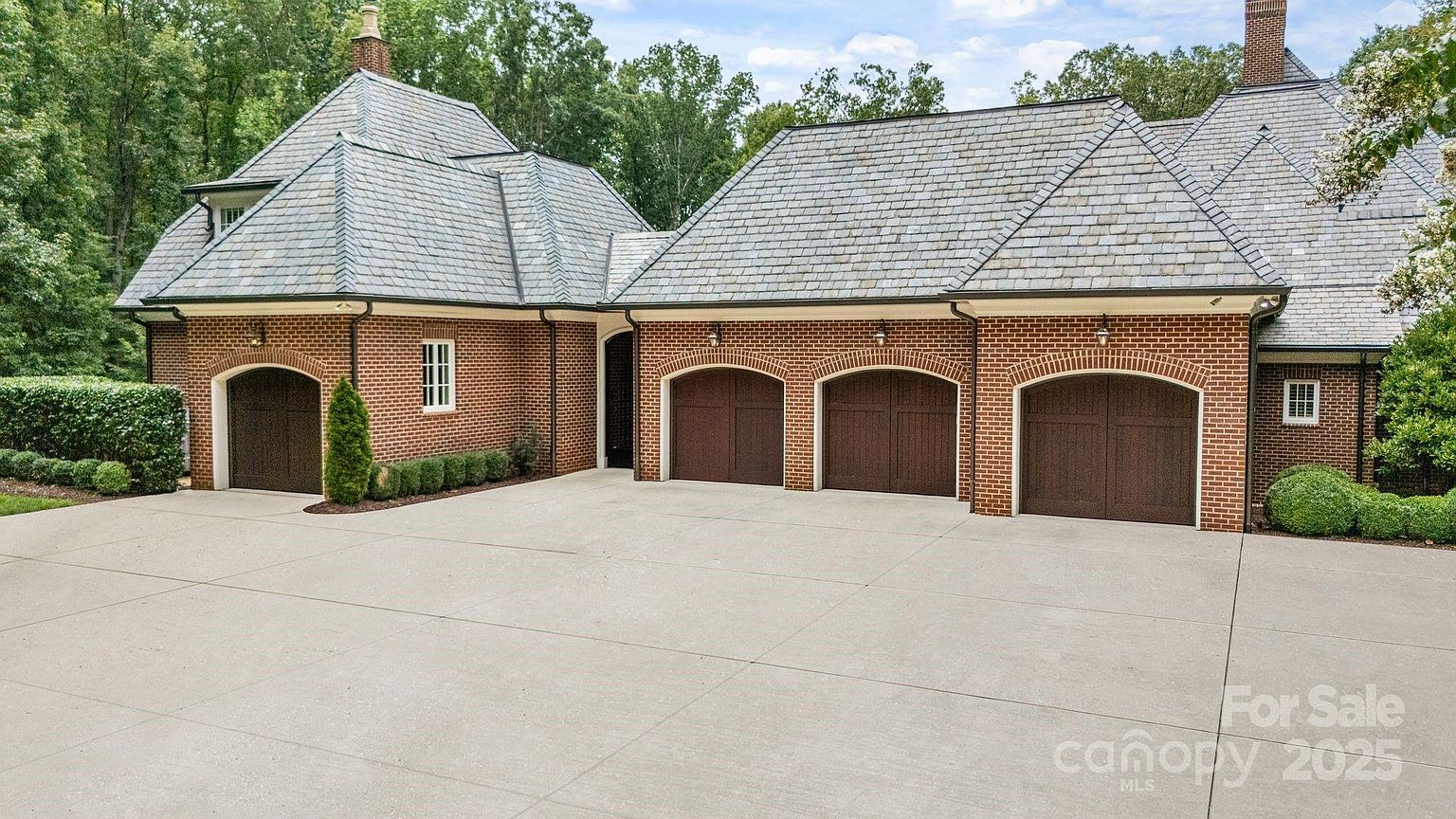 This image showcases a substantial brick-clad garage structure with a multi-car capacity, featuring three arched garage doors and a separate single-car bay to the left. The home is topped with a complex, multi-gabled slate roof and is surrounded by manicured landscaping and mature trees. A wide, clean concrete driveway leads up to the garage, providing a clear and inviting view of the property's exterior architecture.
