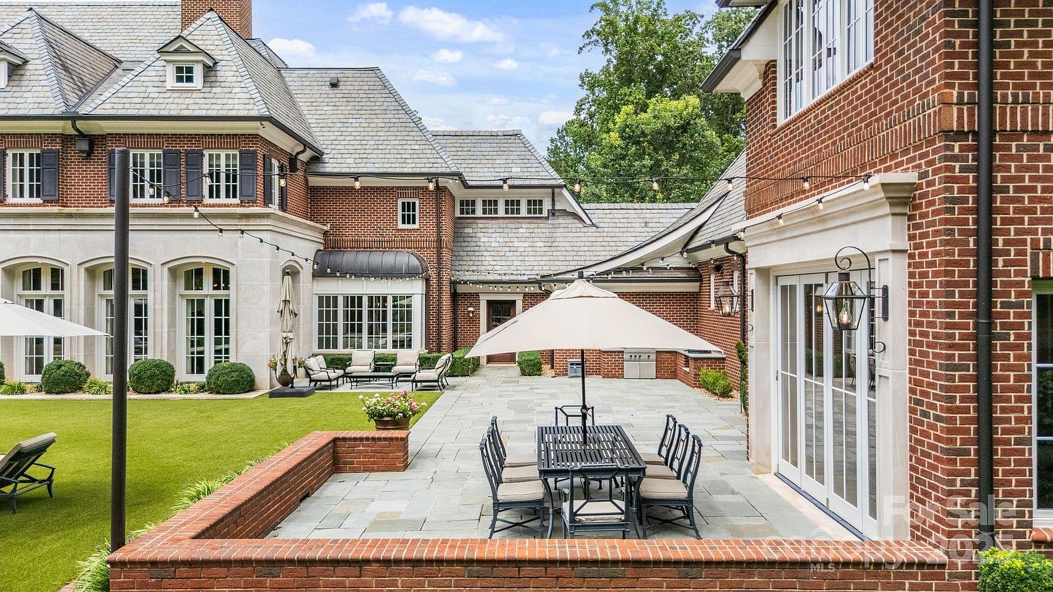 This expansive rear patio features a classic brick and stone exterior design, showcasing a large dining area set beneath a beige umbrella. The space is framed by a low brick retaining wall and overlooks a manicured lawn, with string lights overhead adding a warm, inviting atmosphere. The architecture includes multiple tall windows and glass doors that seamlessly connect the indoor living spaces to the outdoor terrace.