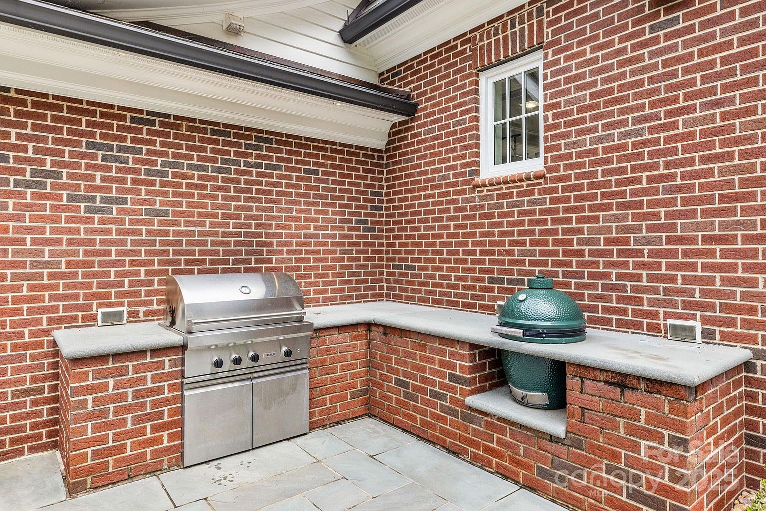 This outdoor kitchen area features a classic red brick wall construction with a stone countertop surface. The space is equipped with a stainless steel built-in grill and a green ceramic kamado-style grill, providing a functional setup for outdoor cooking. The patio is paved with large stone tiles, creating a durable and cohesive look for this exterior entertaining space.