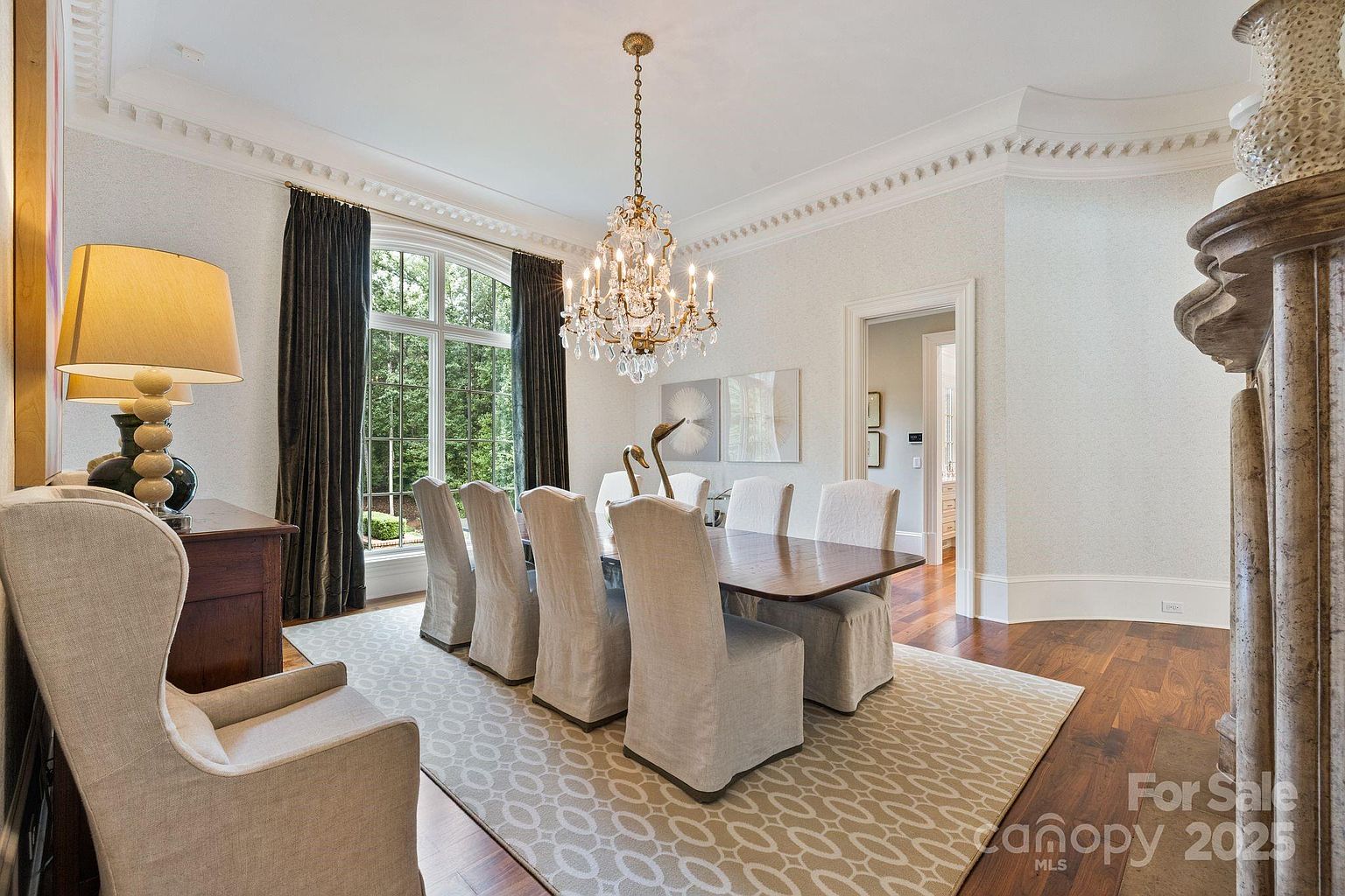 This elegant dining room features a long wooden table surrounded by upholstered chairs, set atop a patterned area rug. A grand crystal chandelier hangs as the focal point above the table, while tall arched windows draped in dark curtains allow natural light to fill the space. The room is finished with detailed crown molding and a stone-clad chimney breast on the right, creating a sophisticated and formal atmosphere.