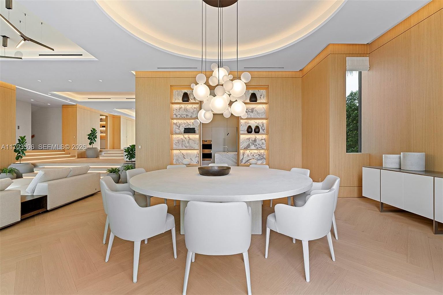 This elegant dining room features a large, circular white table surrounded by modern, minimalist white chairs, positioned beneath a striking, multi-globe chandelier. The space is defined by warm, light-toned wood paneling on the walls and integrated shelving with marble backsplashes, creating a sophisticated and cohesive aesthetic. The open-concept layout seamlessly transitions into a bright living area, emphasizing a sense of luxury and spaciousness.
