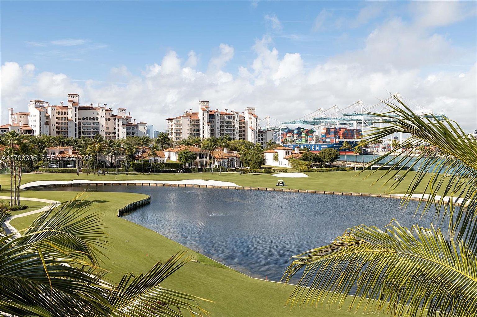 This elevated view captures a serene golf course landscape featuring a central pond and manicured green fairways. In the background, large Mediterranean-style residential buildings stand against a bright blue sky, while a shipping port with cranes is visible in the distance. The foreground is framed by the lush fronds of a palm tree, creating a tropical and tranquil atmosphere.