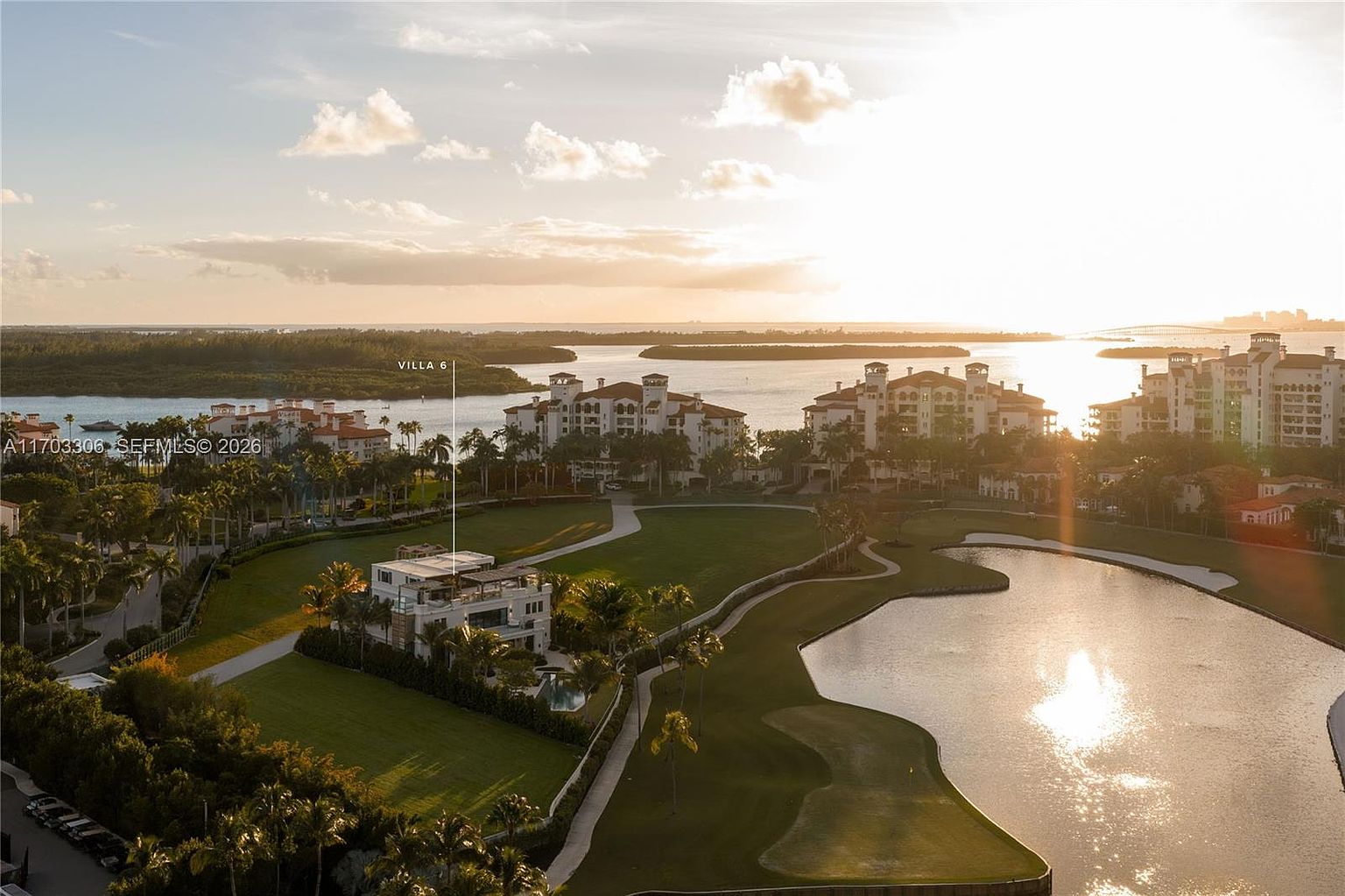 This stunning aerial view captures a luxurious villa nestled within a manicured golf course community, overlooking a serene bay at sunset. The perspective highlights the property's expansive green lawns, private pool, and proximity to the water, emphasizing an exclusive, resort-style lifestyle. The warm golden hour light enhances the tranquil and upscale atmosphere of the estate.